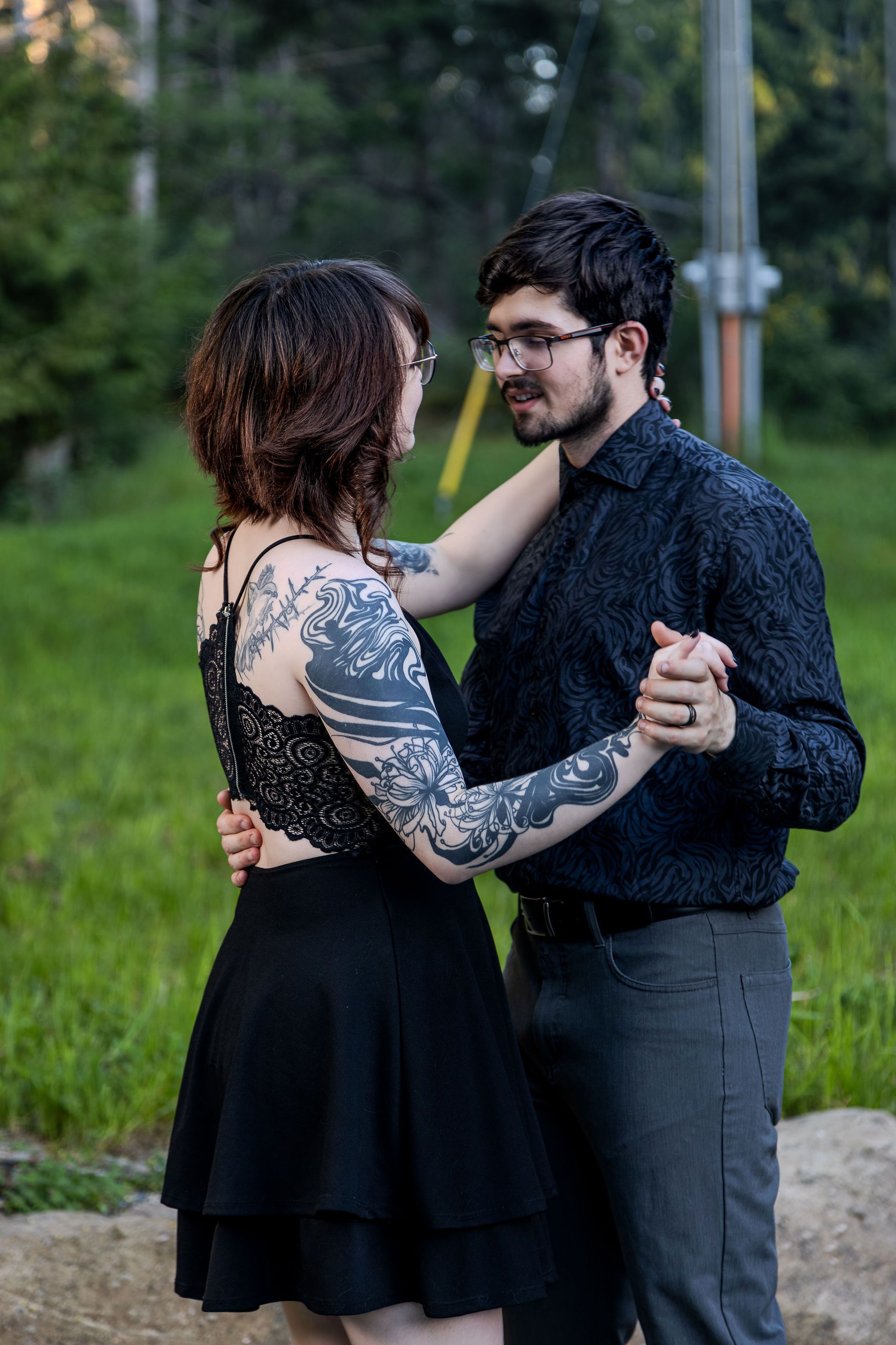 Tattooed couple sharing a slow dance outdoors on rocky path with green meadow behind them.