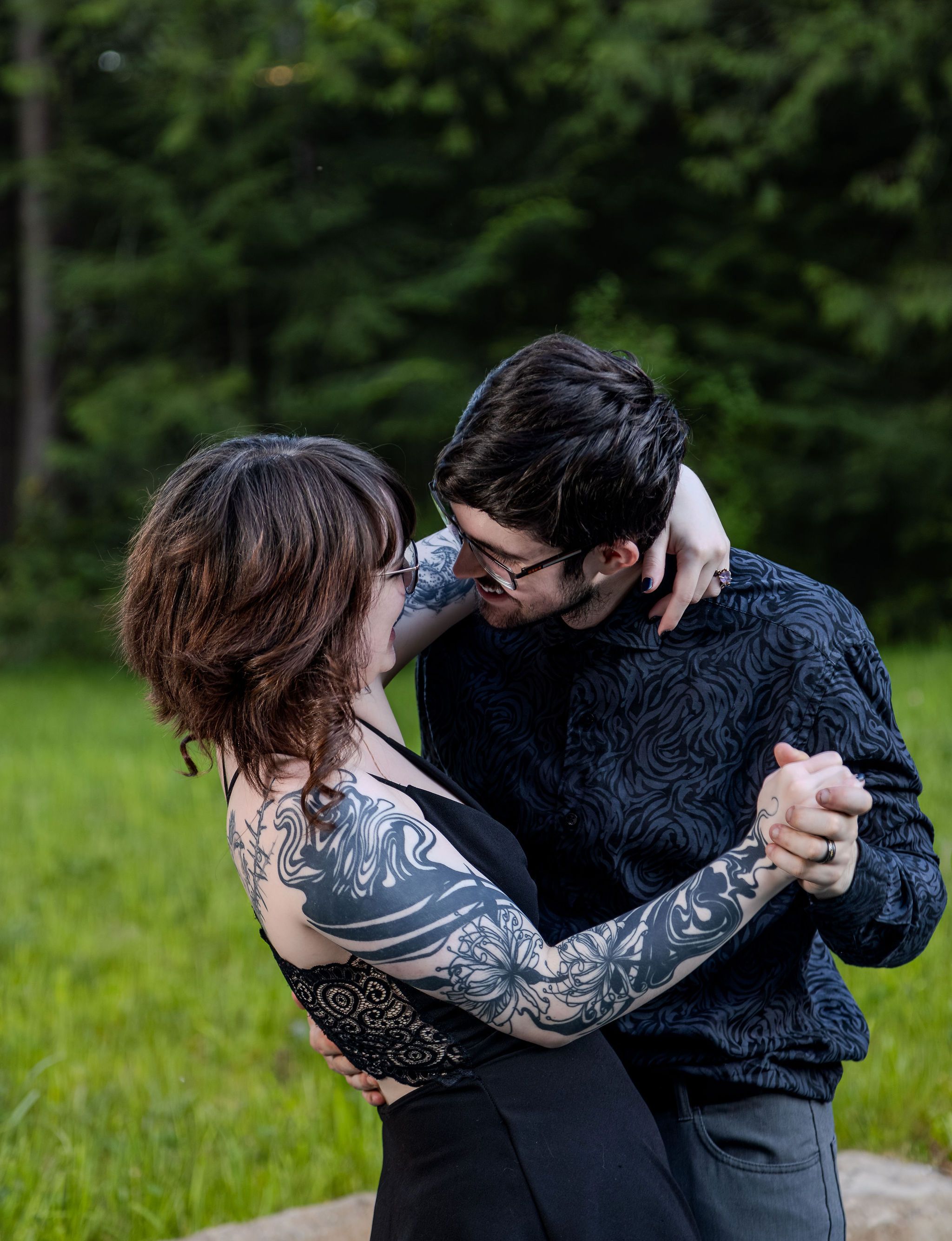 Couple dancing intimately outdoors, woman with tattoo sleeve in black dress embraced by man in dark floral shirt.