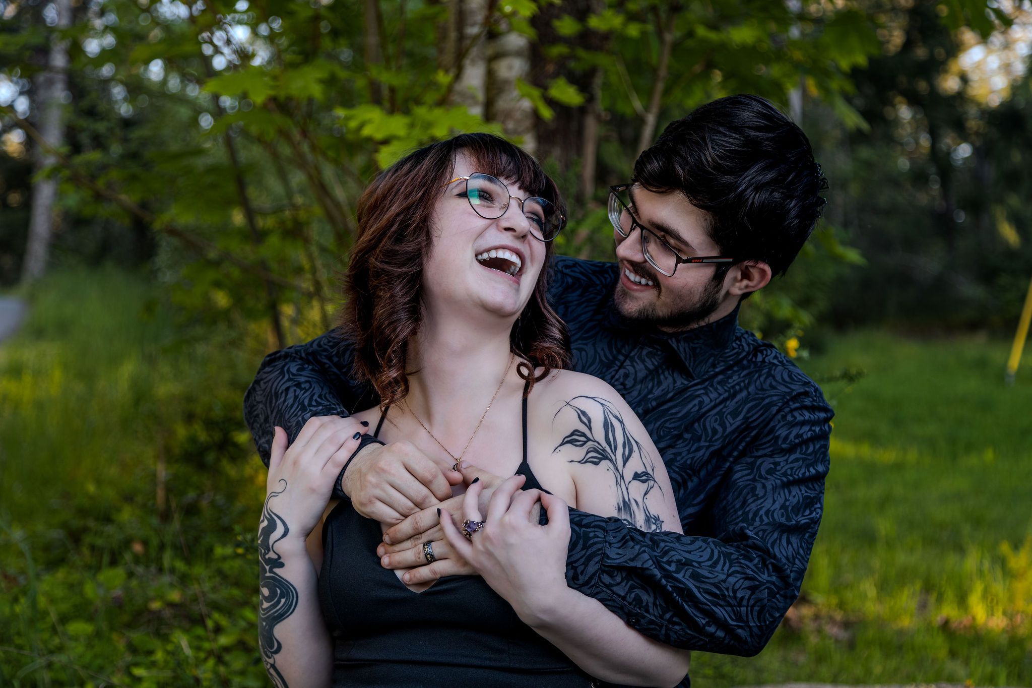 Laughing tattooed couple sharing a joyful embrace outdoors surrounded by lush green trees.
