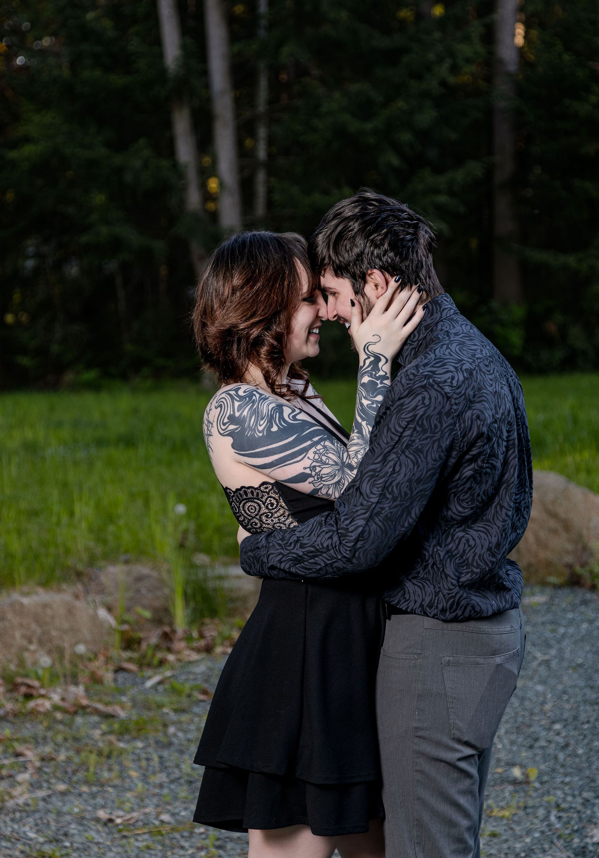 Tattooed couple sharing a romantic nose-to-nose moment outdoors at dusk in forest setting.