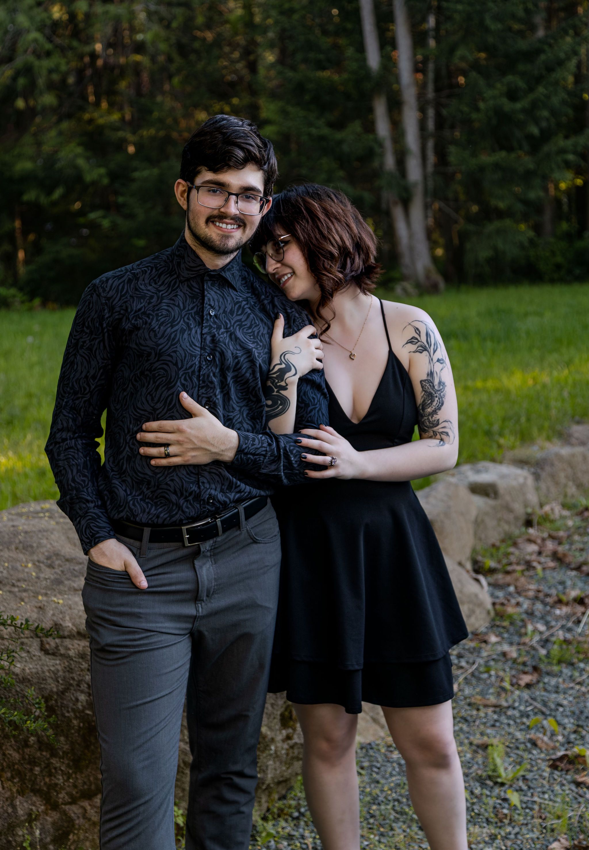 Smiling tattooed couple posing casually on rocks with lush green forest background.