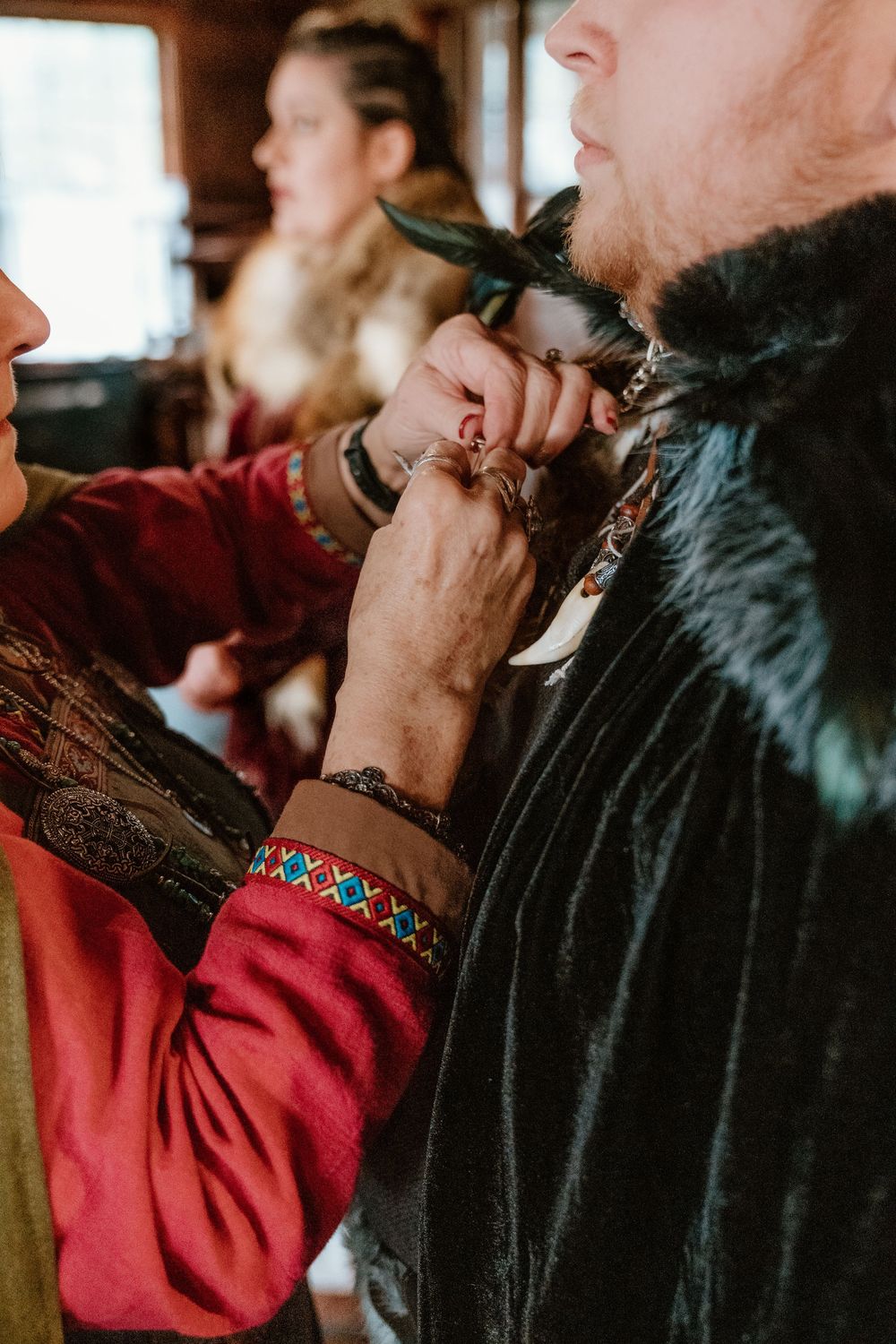Person in Viking attire adjusting another's braided beard, with woman in medieval costume visible in background.
