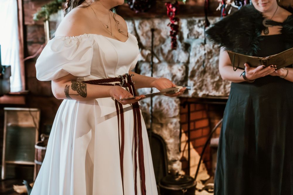 Bride in white off-shoulder gown with brown belt reads vows at rustic indoor wedding ceremony.