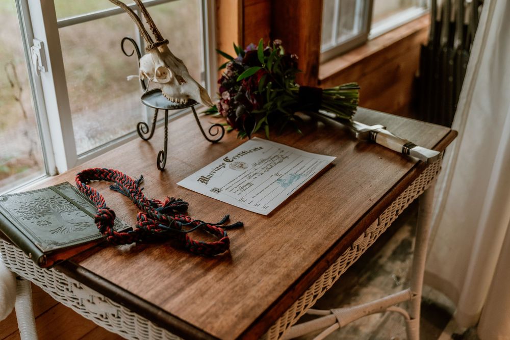 Rustic wooden desk with marriage certificate, rosary beads, open book, and floral bouquet near a window.