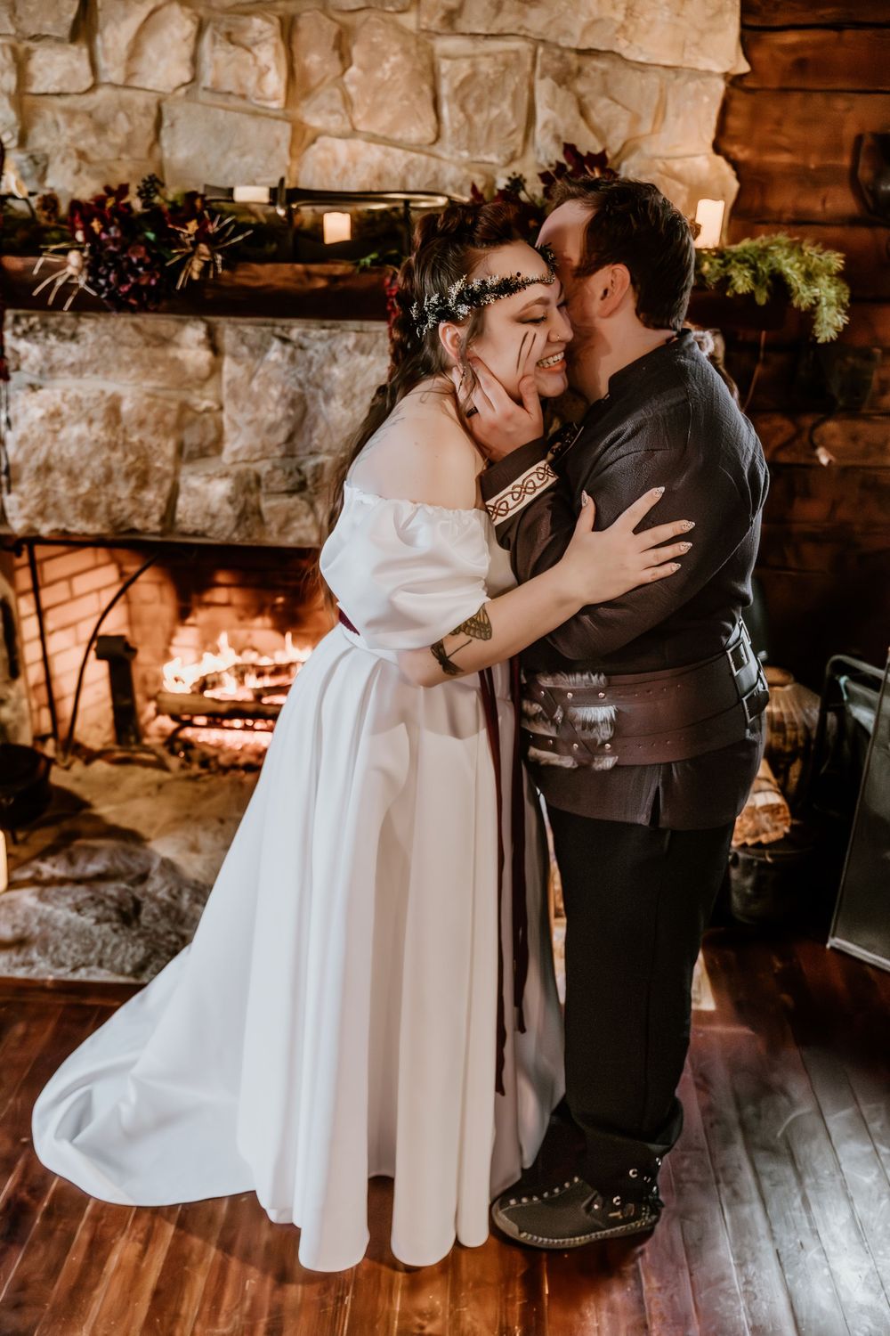 A bride and groom share a romantic kiss in front of a stone fireplace, she in a white gown and floral crown.