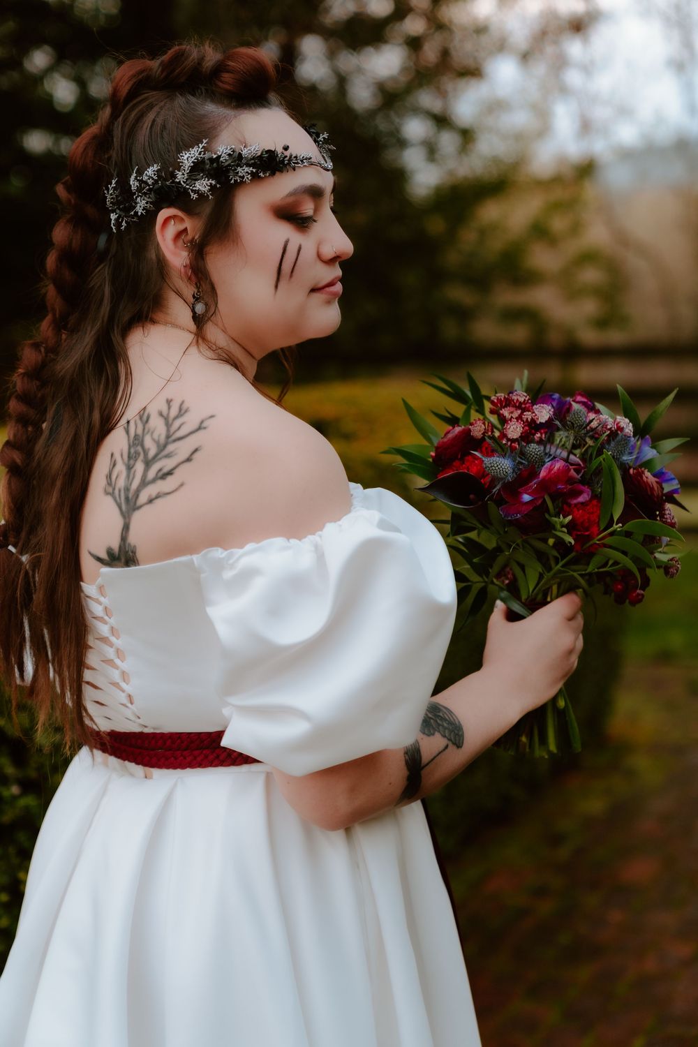 A plus-size bride in an off-shoulder white gown with red belt holds a dark floral bouquet, featuring tattoos and dramatic makeup.
