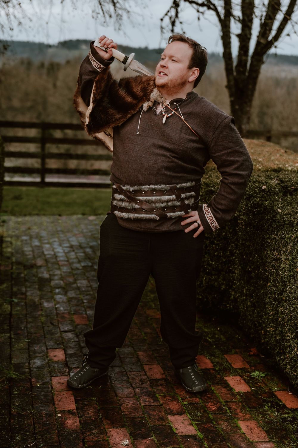 Man in Viking costume holding an axe aloft outdoors on a brick patio, wearing fur and medieval clothing.