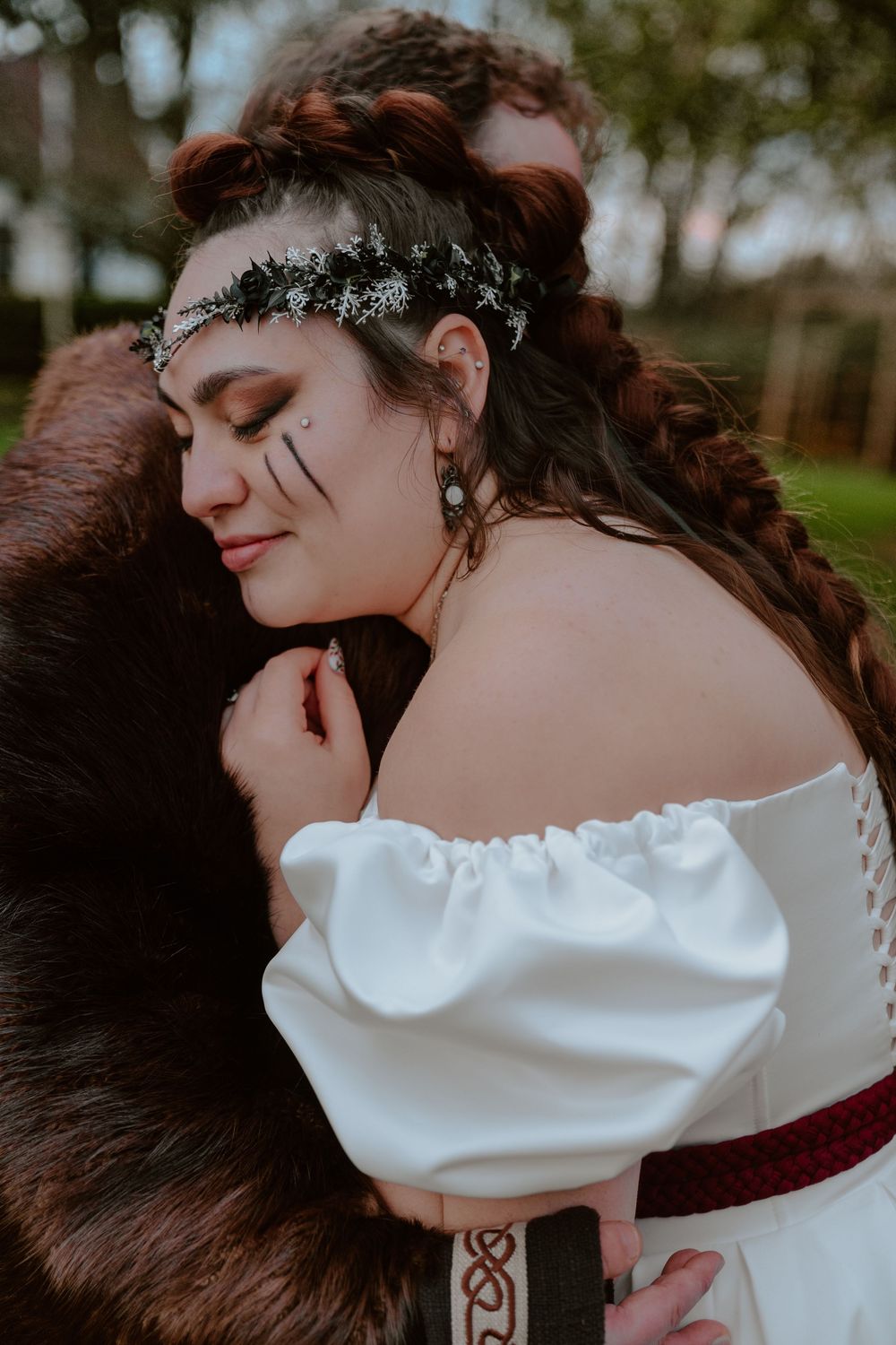 Woman in white off-shoulder dress with floral crown and face art, embraced in a fur wrap outdoors.