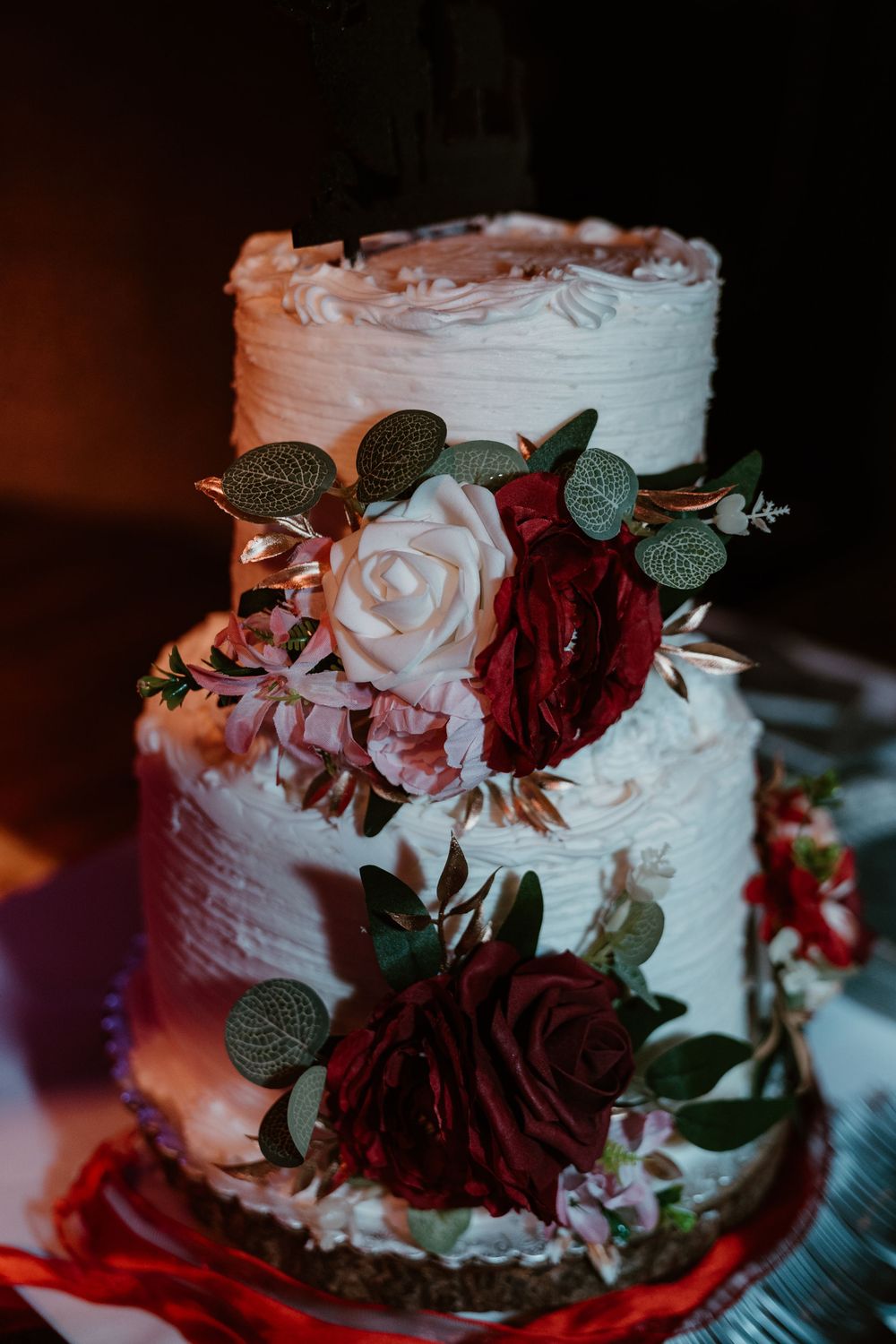 Two-tier white textured wedding cake adorned with burgundy and blush roses, eucalyptus, and gold leaves.