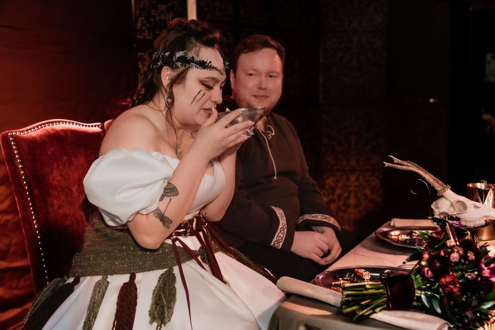 Bride and groom in gothic wedding attire sit at decorated table, bride emotional, groom smiling softly.