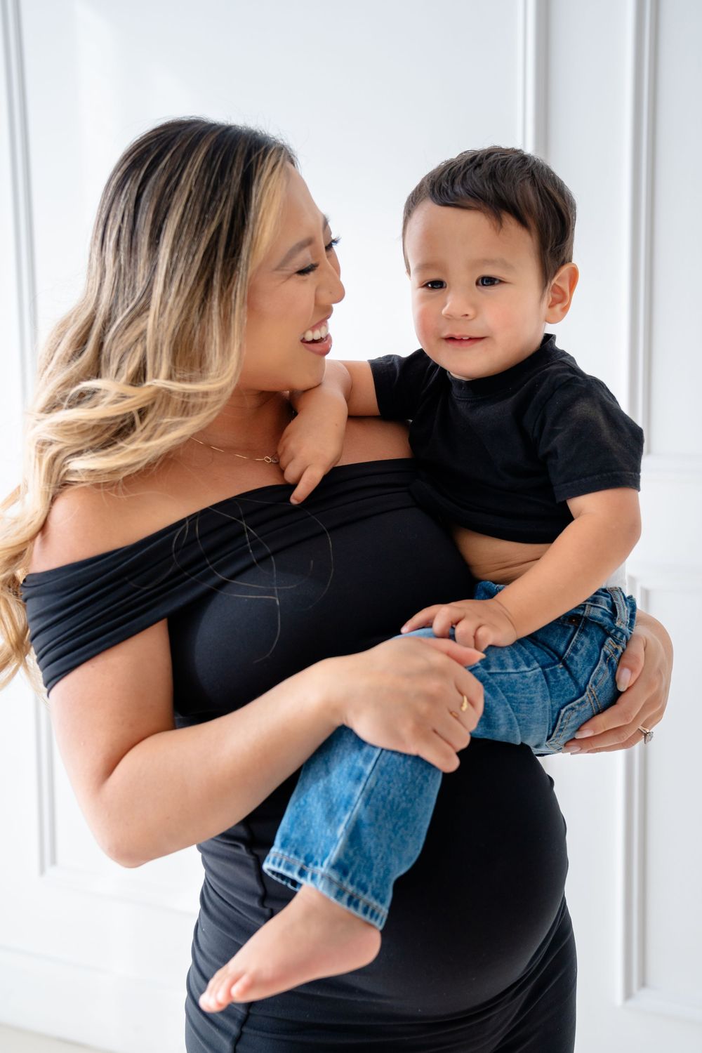 Mother in black shirt holds and hugs playful toddler in casual wear against white curtained backdrop.