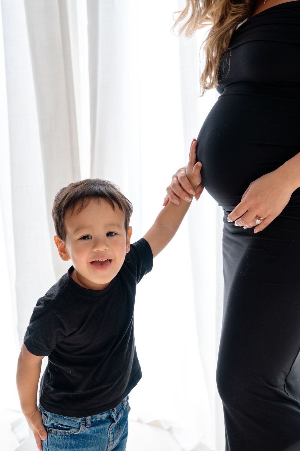 Toddler in black t-shirt and jeans plays and smiles against white curtain background in photo series.