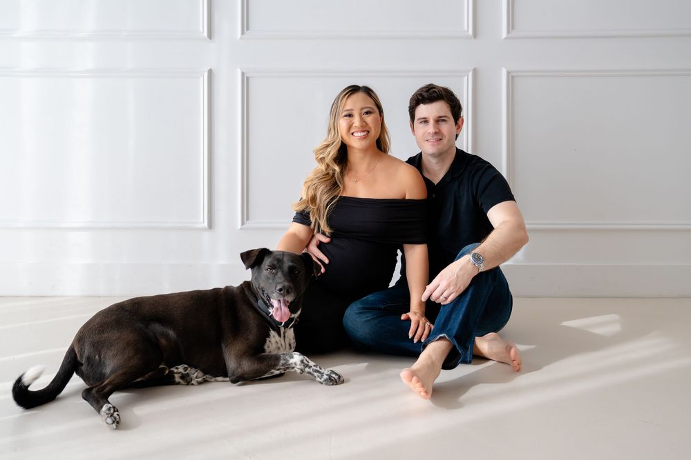 A couple in black outfits poses on a white floor with their large black and white dog in a bright studio setting.