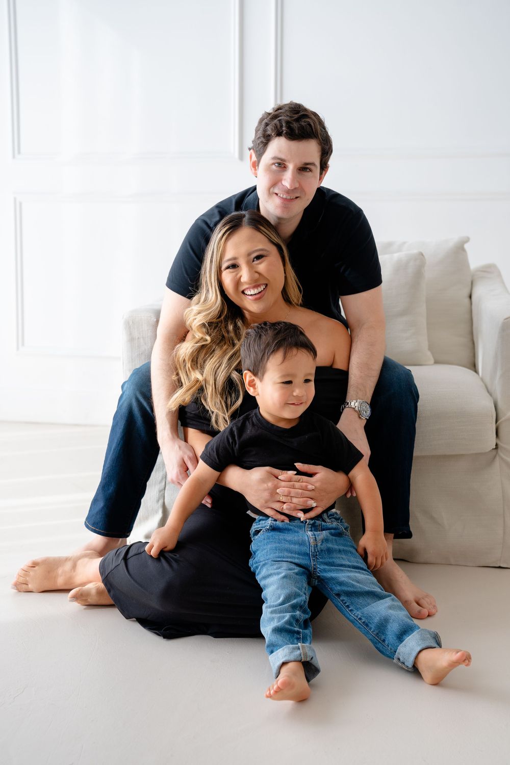A family in black and denim sits casually together on a white floor in a bright studio setting.