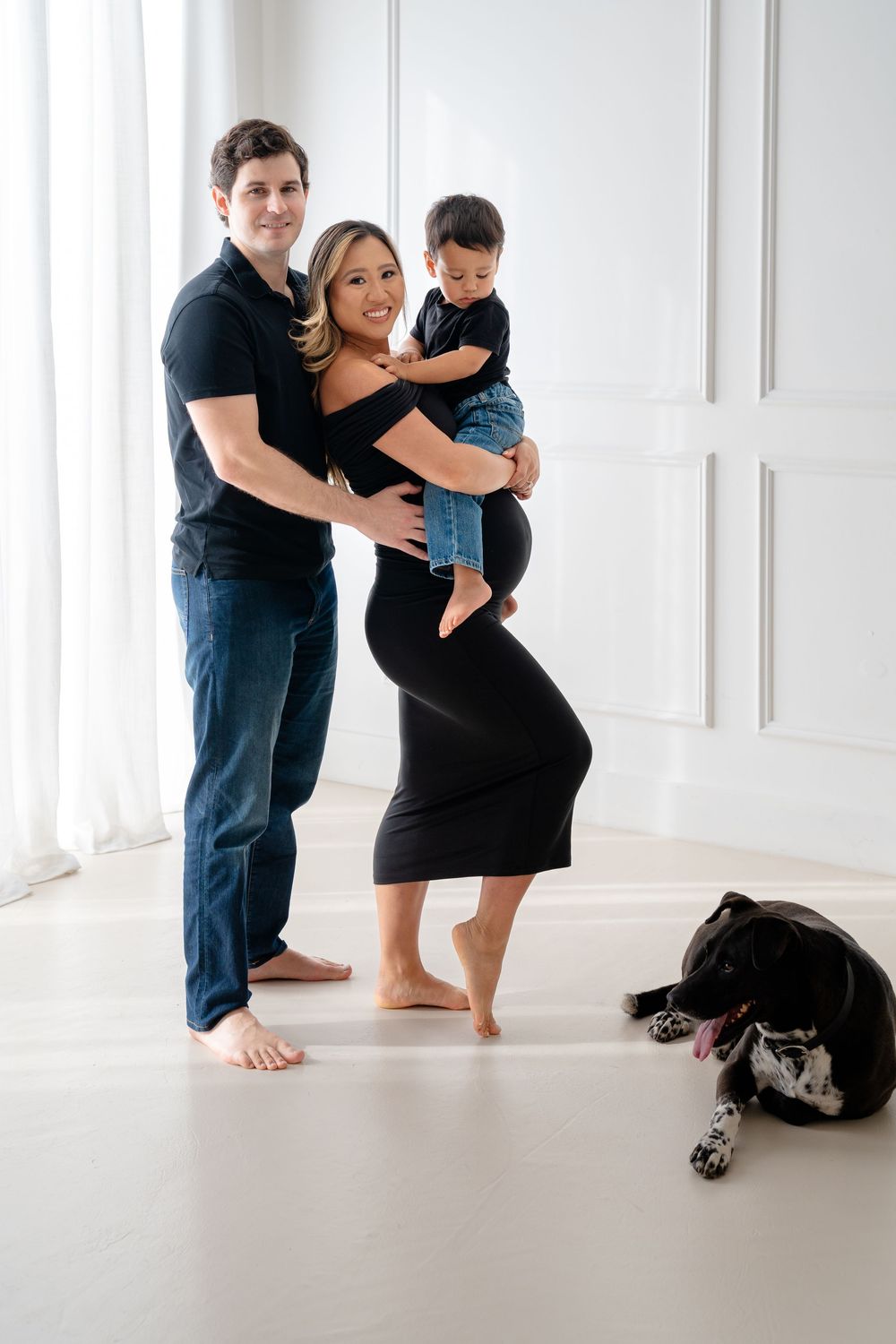 A family poses together in a minimalist white studio setting while their black dog rests nearby.