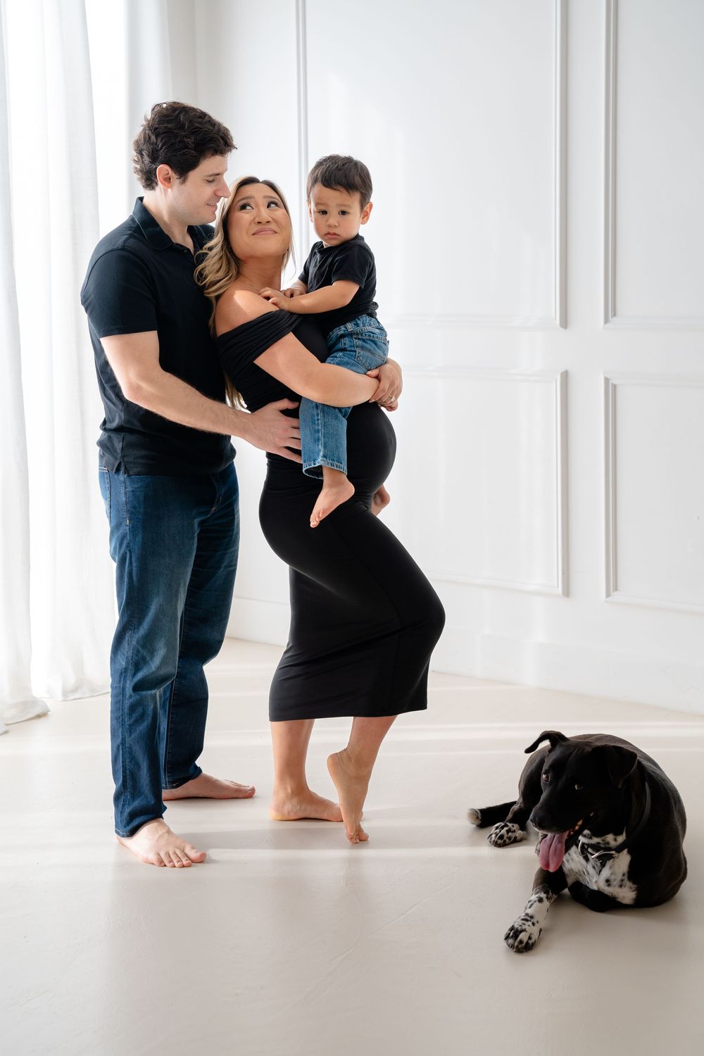 A family shares tender moments in a bright white room with their black dog resting nearby.