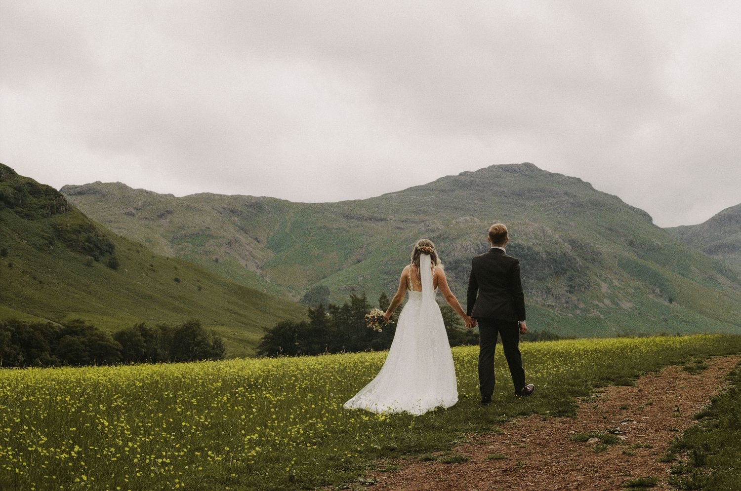 A couple in wedding attire stands on a scenic mountain path surrounded by green hills and wildflowers in Scotland.