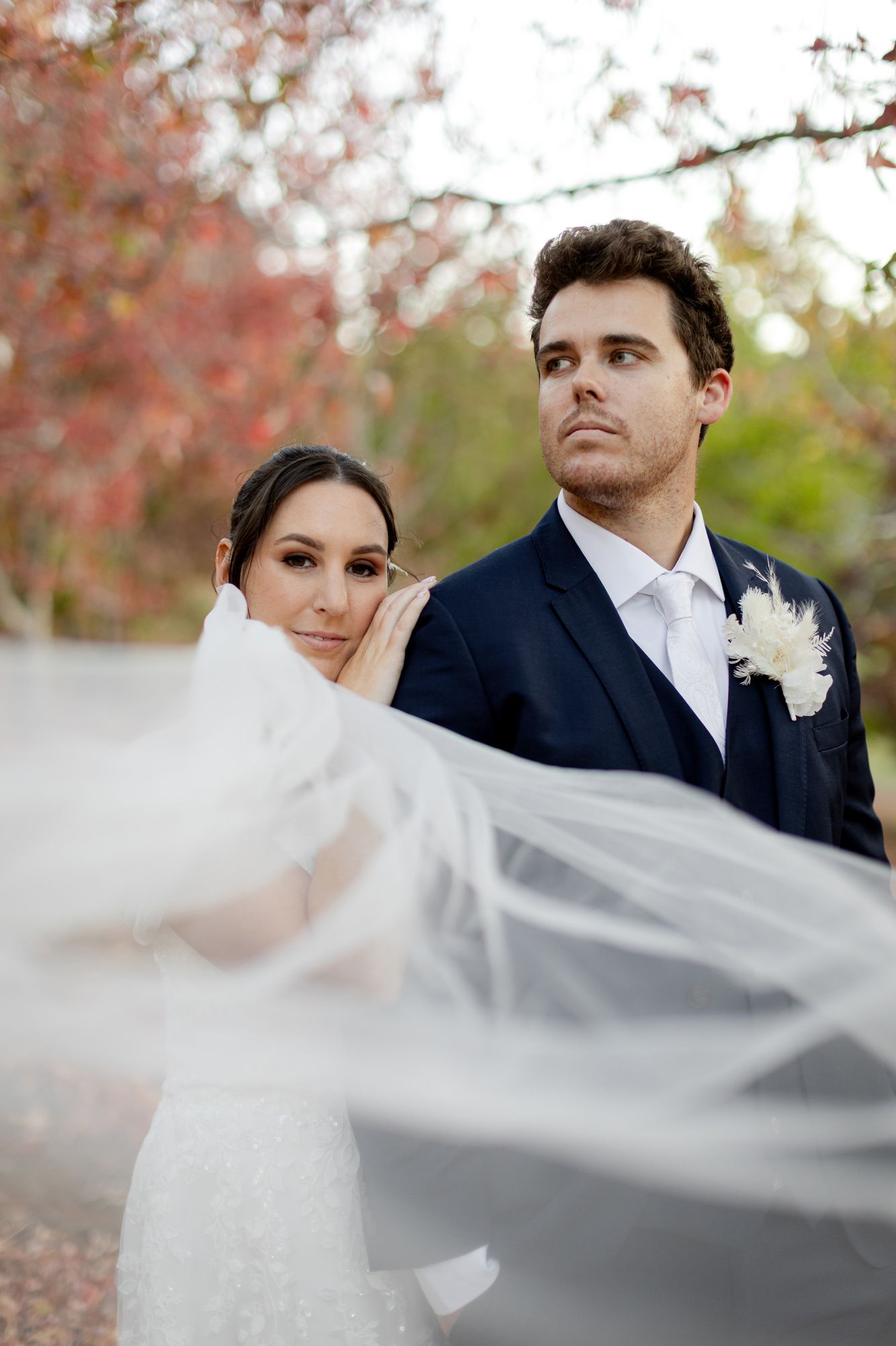 A couple embraces outdoors in wedding attire, with the bride in white lace and groom in navy suit on a sunny garden day.