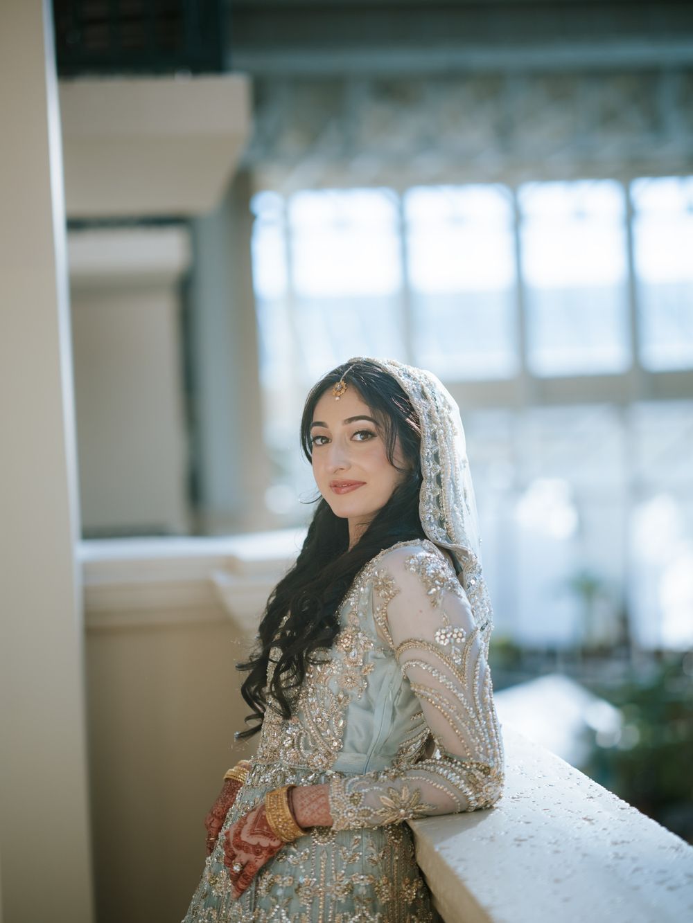 A bride in an elegant white lace wedding dress with long sleeves and veil poses near a window in soft natural light.