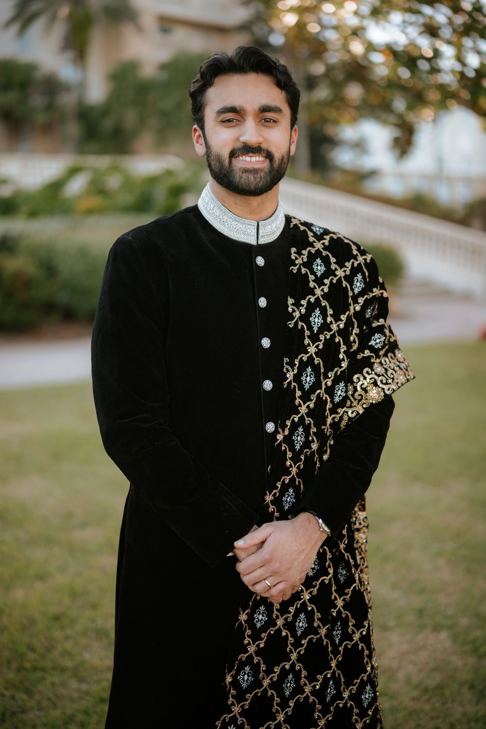 A person wearing a black traditional sherwani with intricate gold embroidery details stands outdoors in a natural setting.