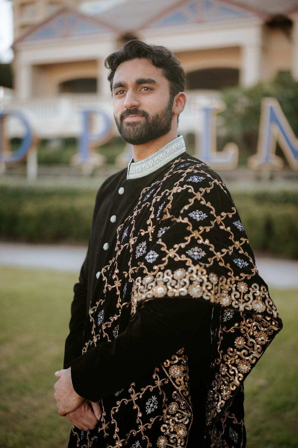 Elegant South Asian groom wearing traditional black and gold embroidered sherwani standing outdoors.