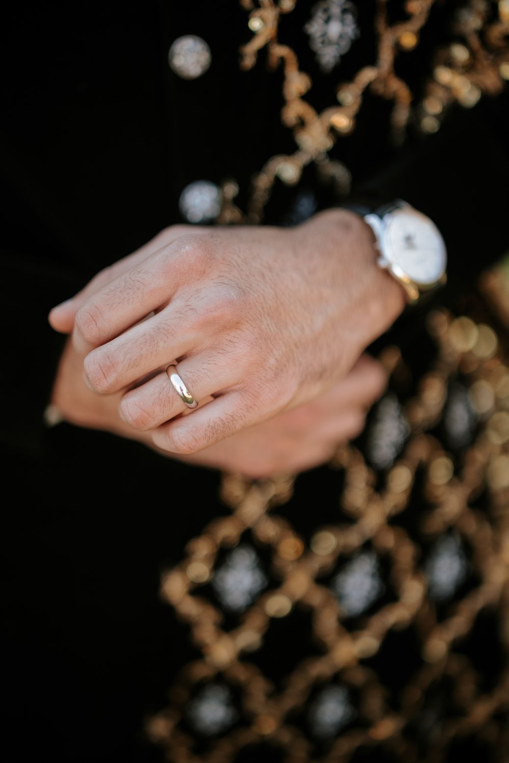 Close-up of a hand wearing a delicate gold engagement ring against a dark background with bokeh lights.
