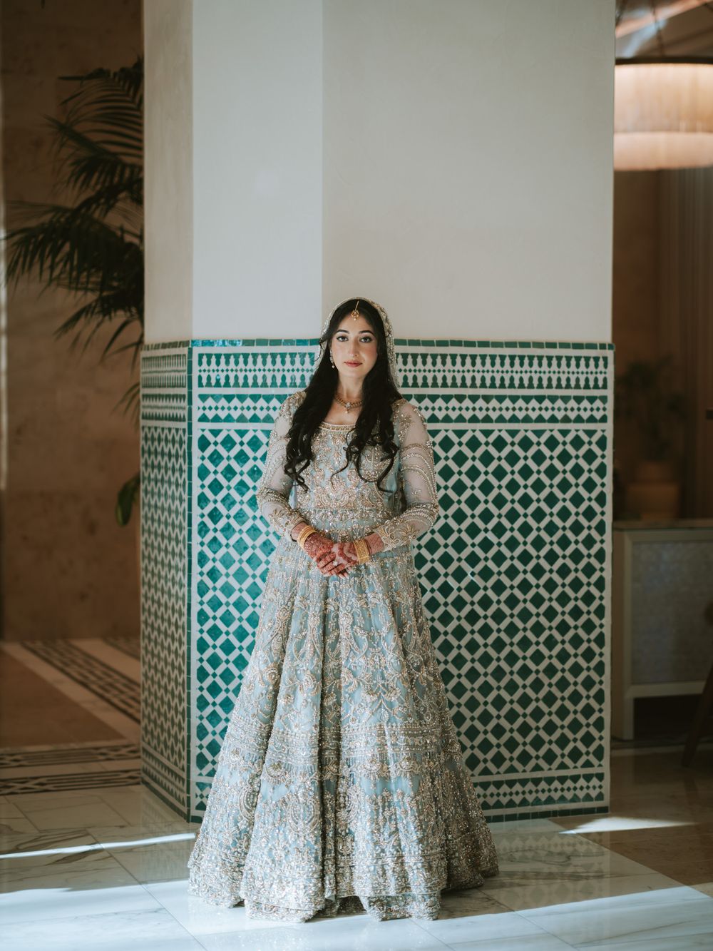 A person in a floral print dress poses against a turquoise and white geometric tiled wall in an elegant interior setting.