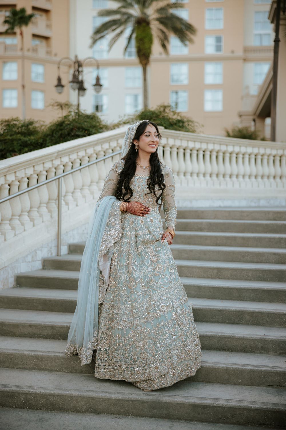 A person in an ornate silver embellished gown poses elegantly on white stone steps near a palm tree at sunset.
