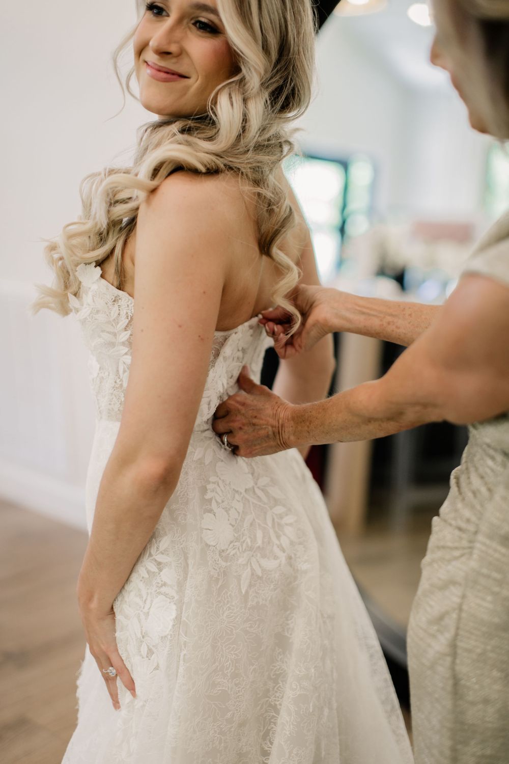 Getting ready for the wedding day, a bride in a lace dress gets help with final adjustments.