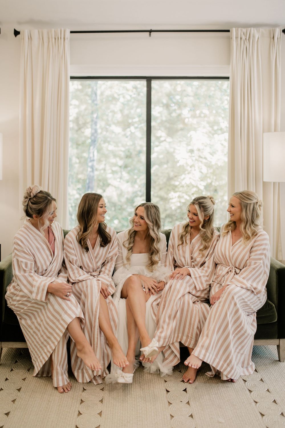 Bridesmaids in striped robes relax together on a white couch during wedding preparations.