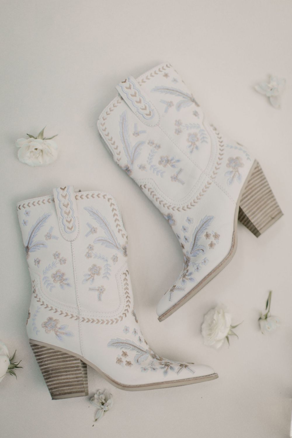 Delicate white cowboy boots with floral embroidery arranged on light background.