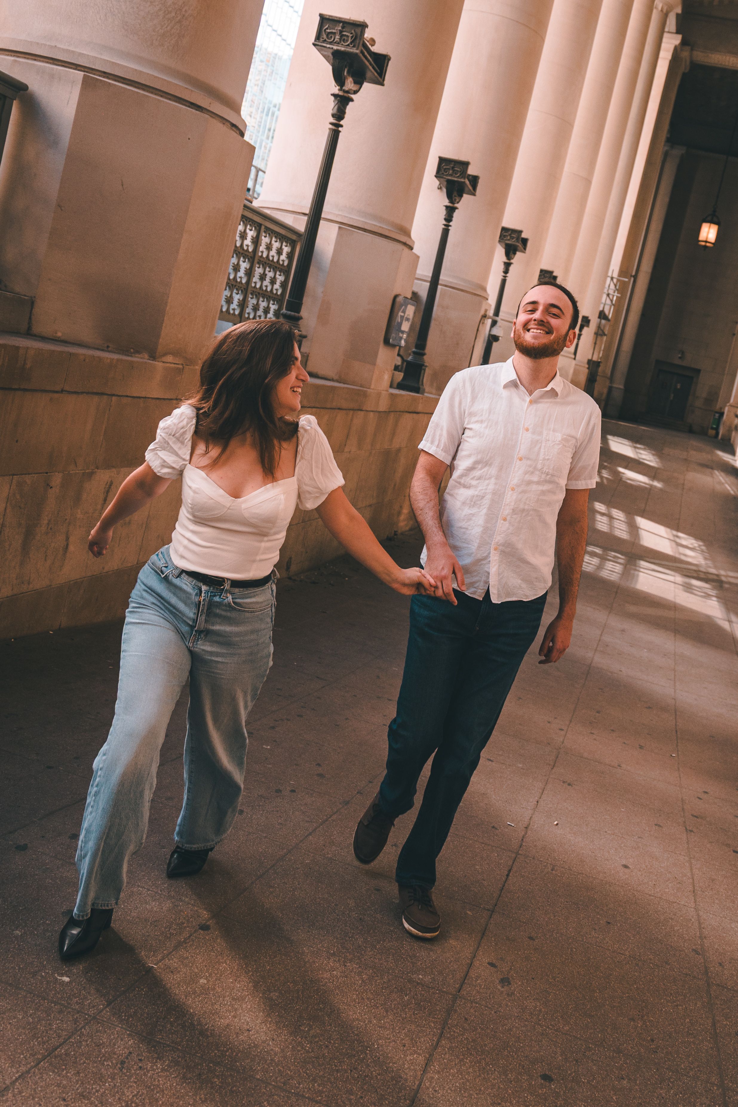 A couple walking through a sunlit colonnade with classical architectural columns creating dramatic shadows.