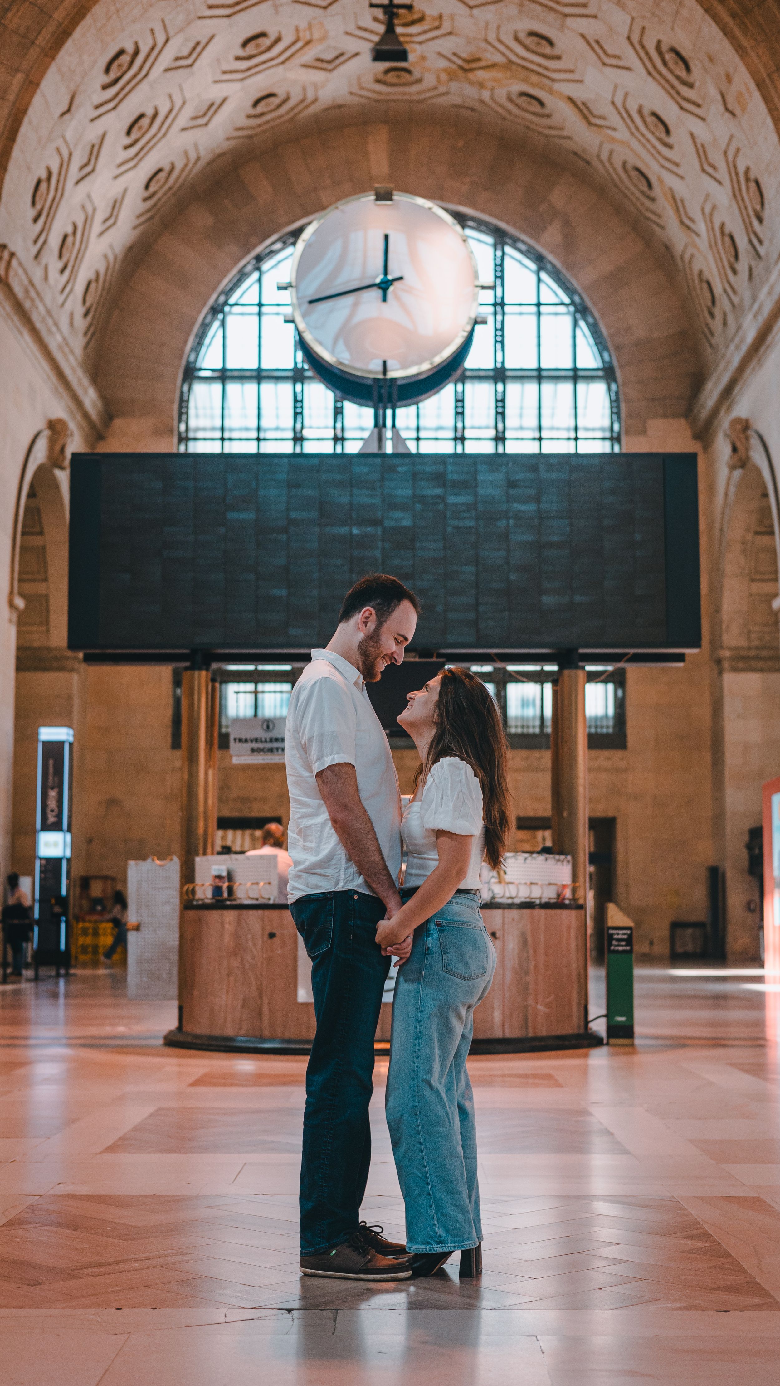 Two people stand in conversation at Ellis Island's historic Great Hall with its vaulted ceiling and arched windows.