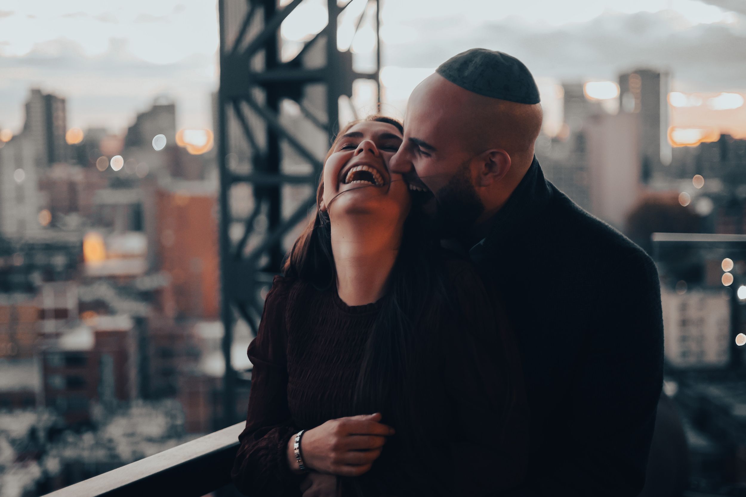A joyful candid moment shared between two people laughing together on a rooftop with city buildings in the background.