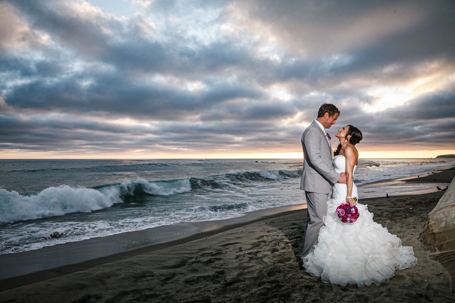A couple embraces on a dramatic beach at sunset with crashing waves and cloudy skies in the background.
