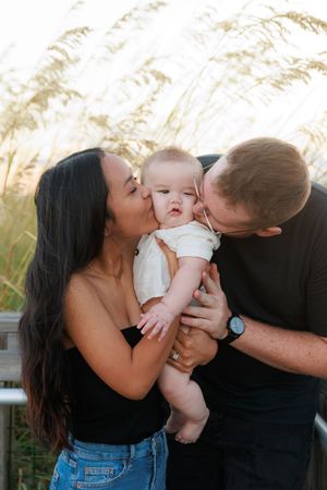 A couple kisses their baby in a series of three tender outdoor photos with tall grass in the background.