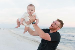A person lifts a baby up in the air while playing at the beach during sunset.