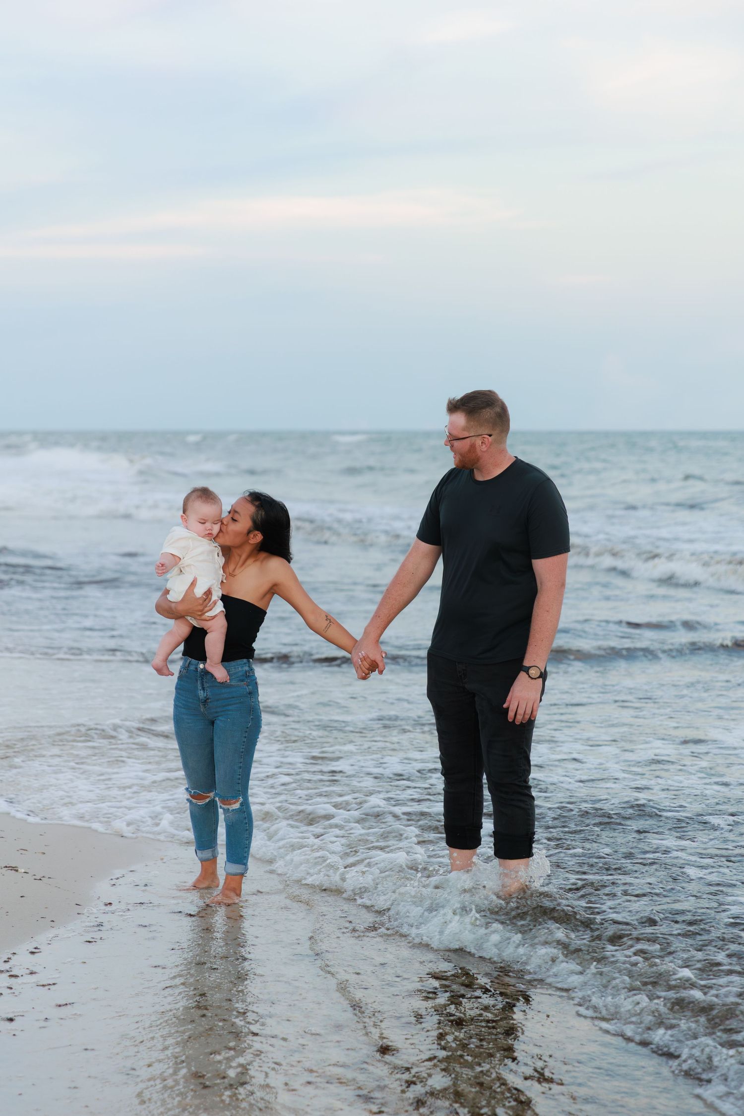 A family stands together holding hands at the edge of the ocean waves during sunset on a peaceful beach.