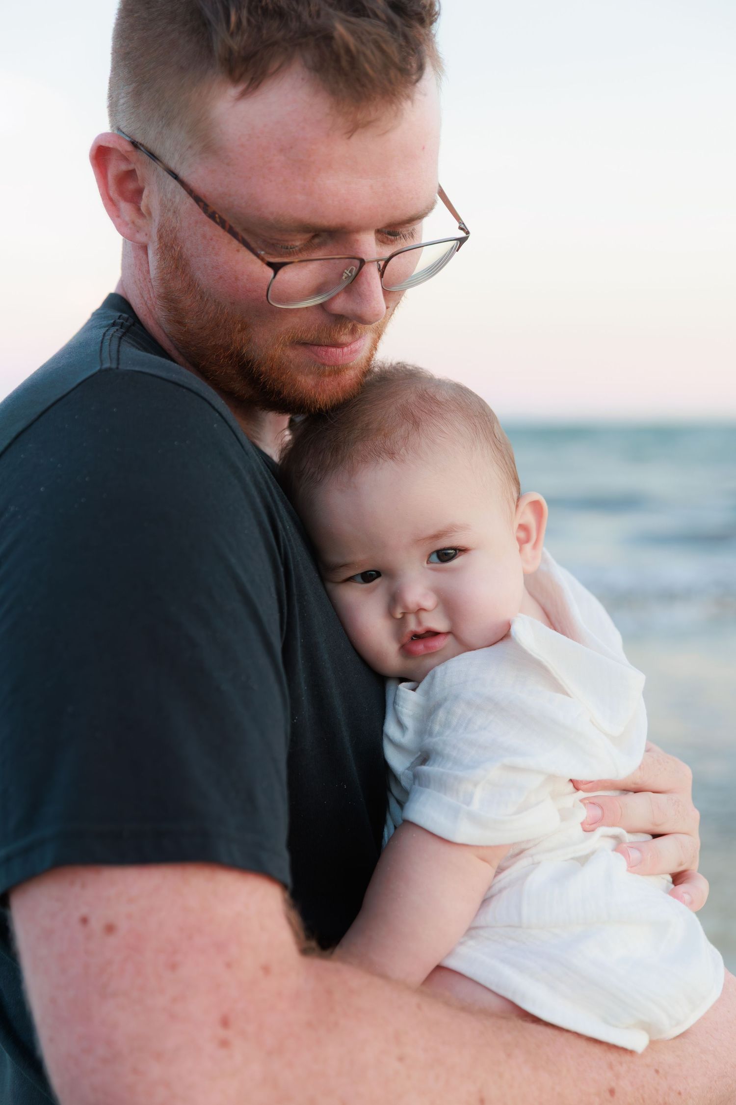 A tender moment captured at the beach as a parent in black shirt embraces a baby in white clothing at sunset.