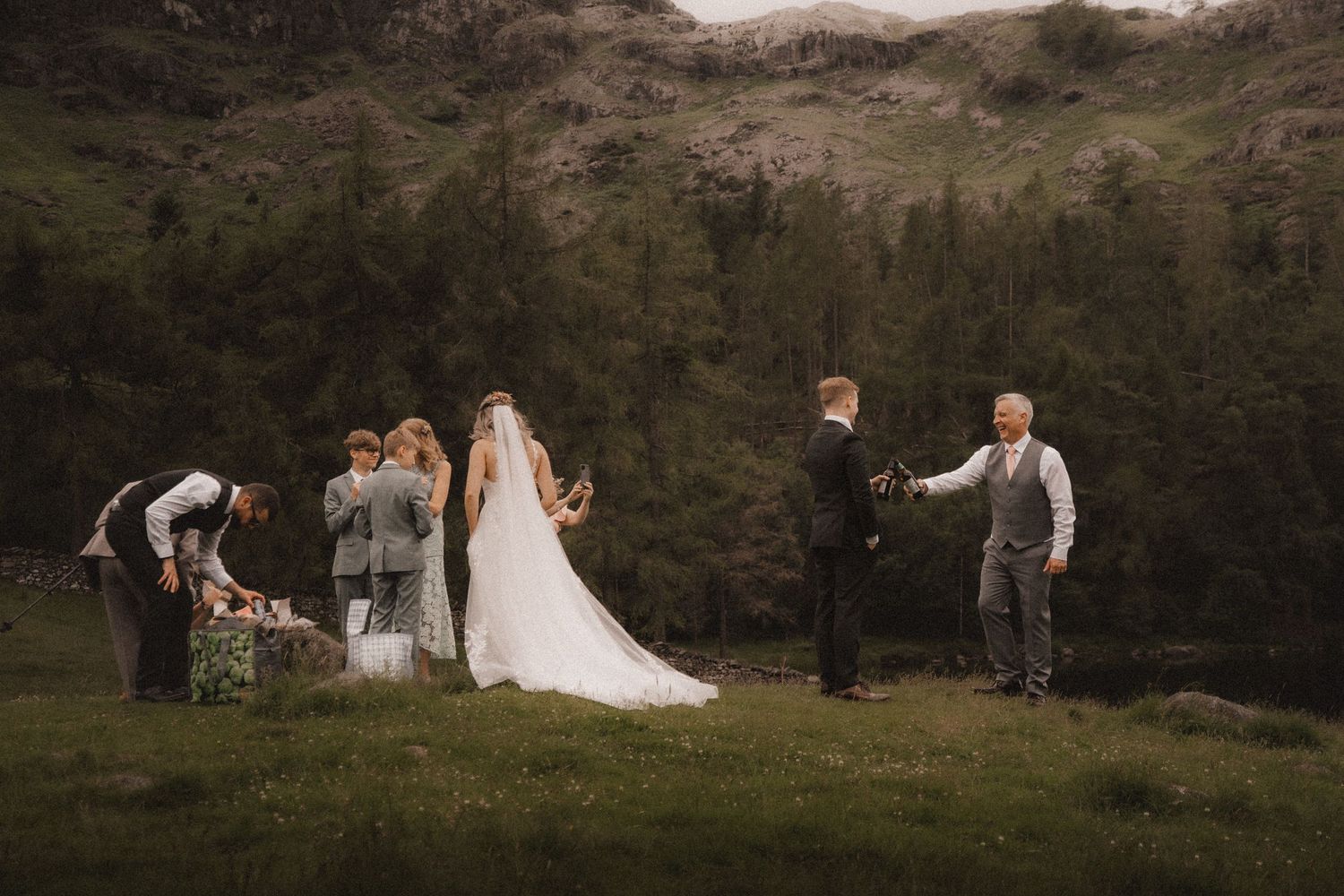 A romantic wedding couple and their guests walk across a scenic hillside meadow at golden hour.