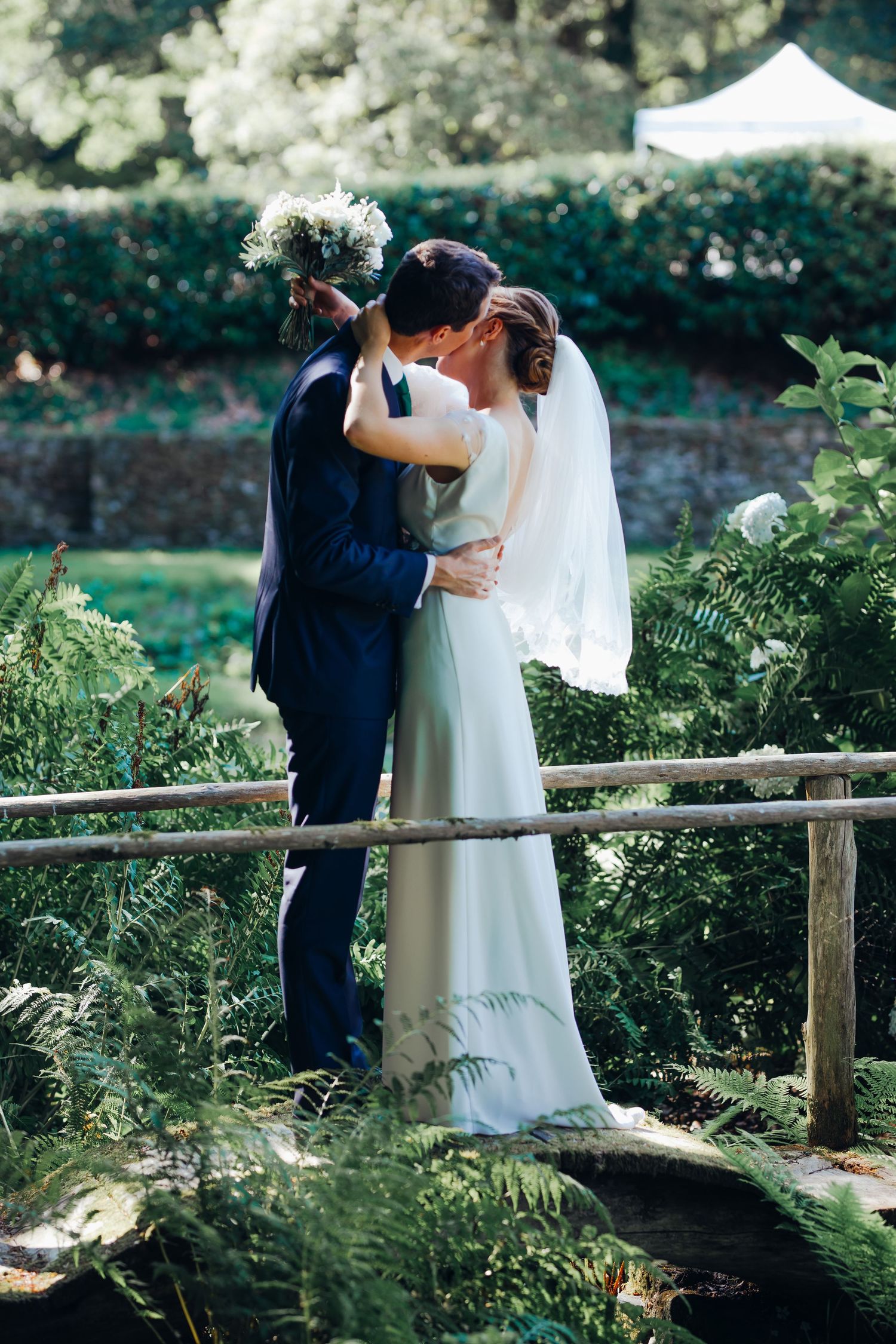 Bride and groom sharing a kiss on a rustic wooden bridge surrounded by lush green ferns.
