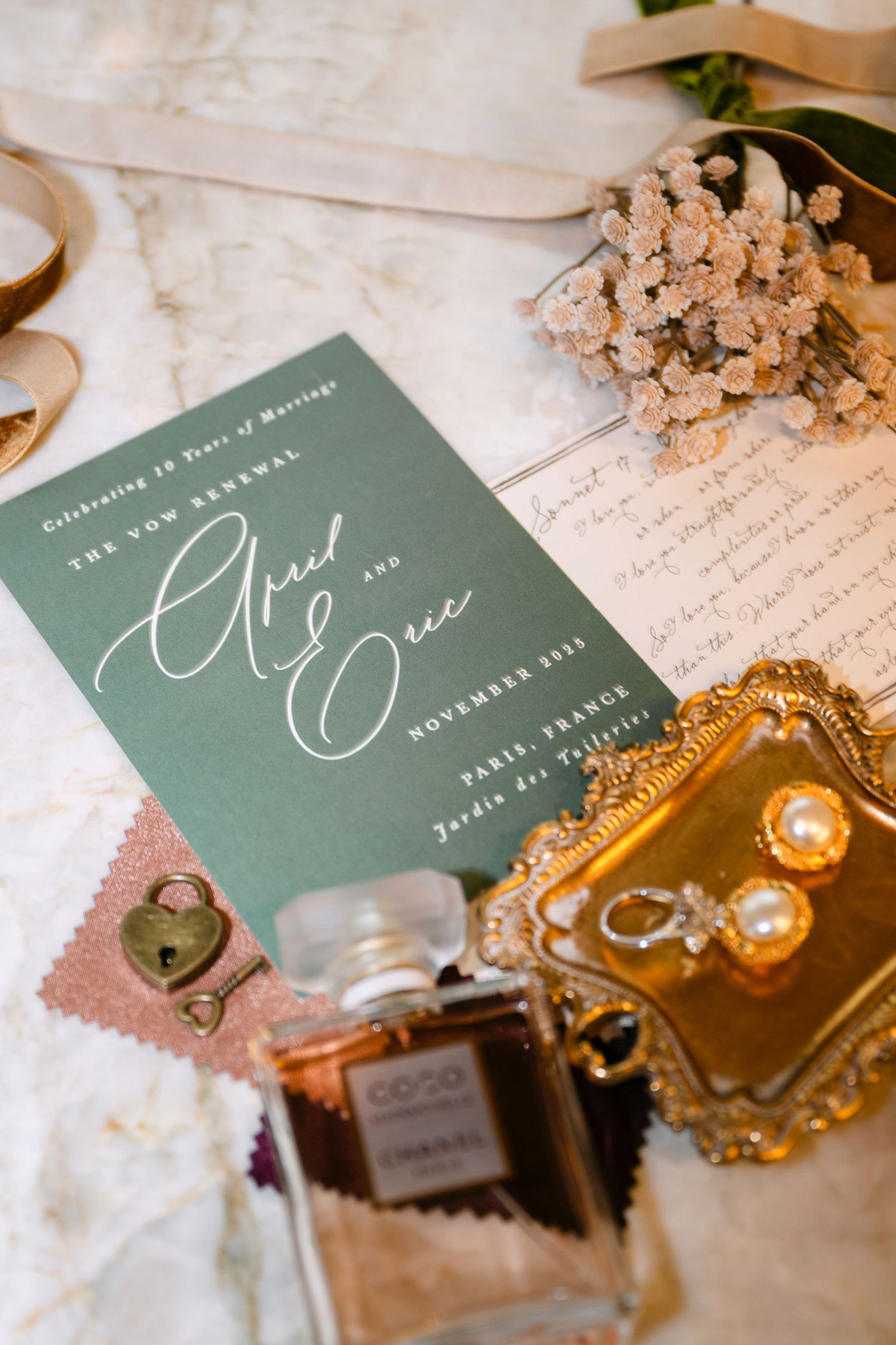 Wedding invitation in sage green with elegant calligraphy displayed alongside perfume bottle and vintage gold jewelry tray.