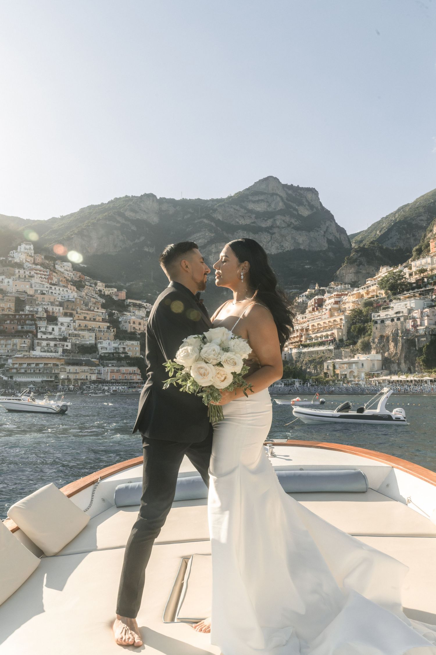 Wedding couple embracing on boat with Positano's picturesque coastal backdrop.