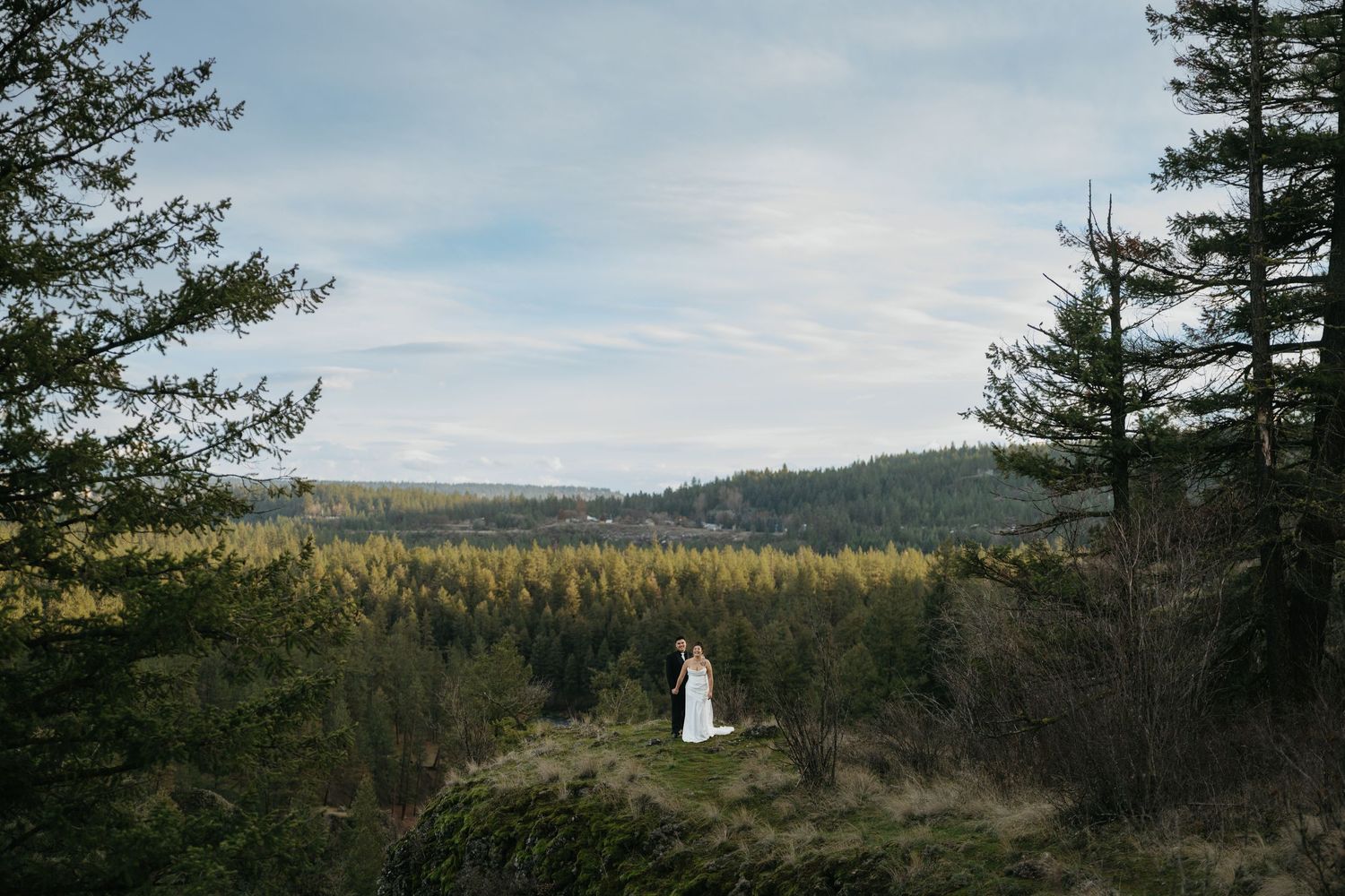 A couple stands on a scenic overlook surrounded by evergreen forest and rolling hills under a cloudy sky.