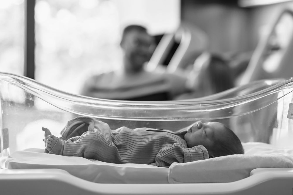Newborn baby resting in clear hospital bassinet with parents in background.
