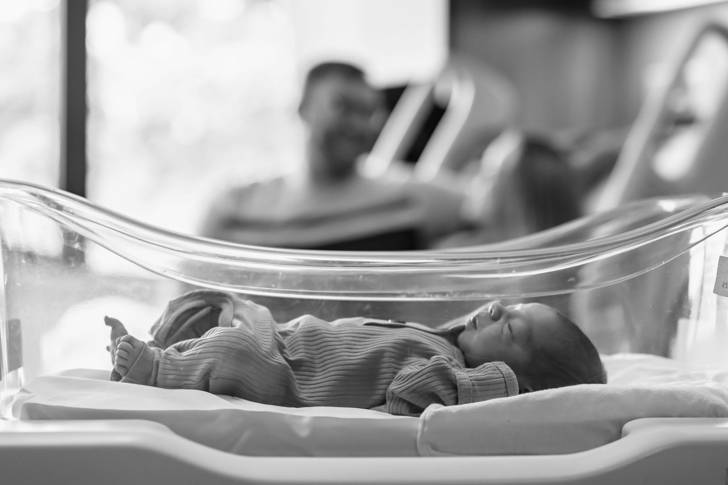 Newborn baby resting in clear hospital bassinet with parents in background.