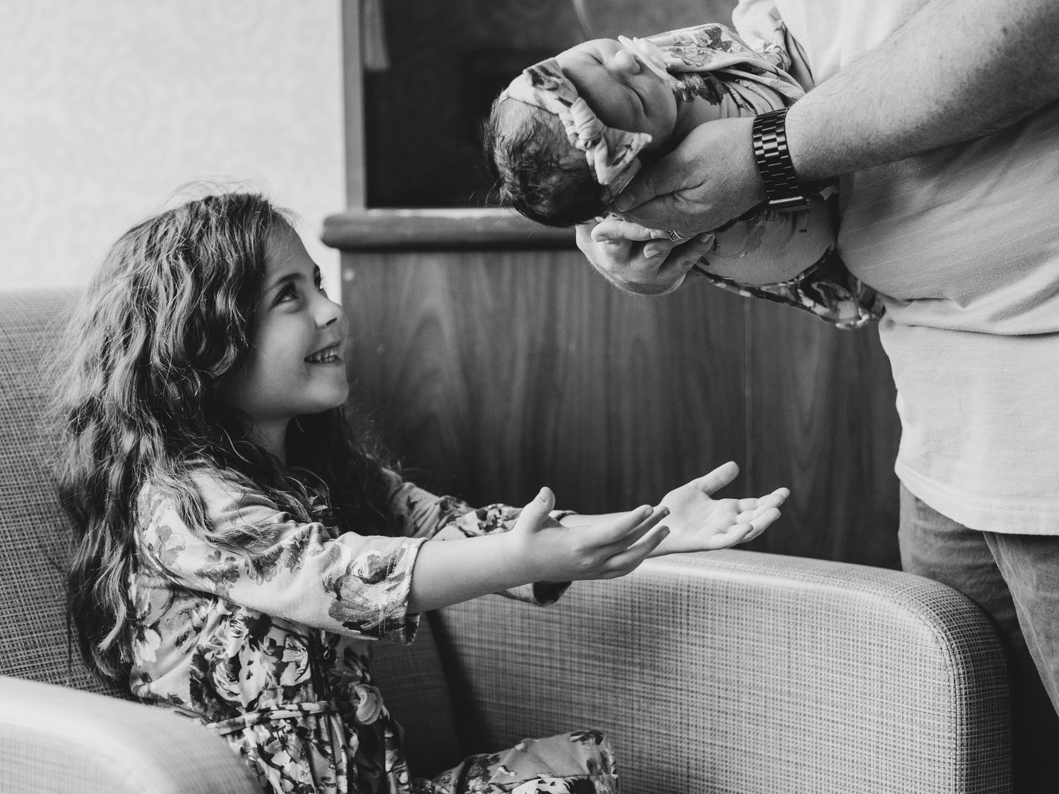 Black and white photo of parent handing baby to excited child on couch.