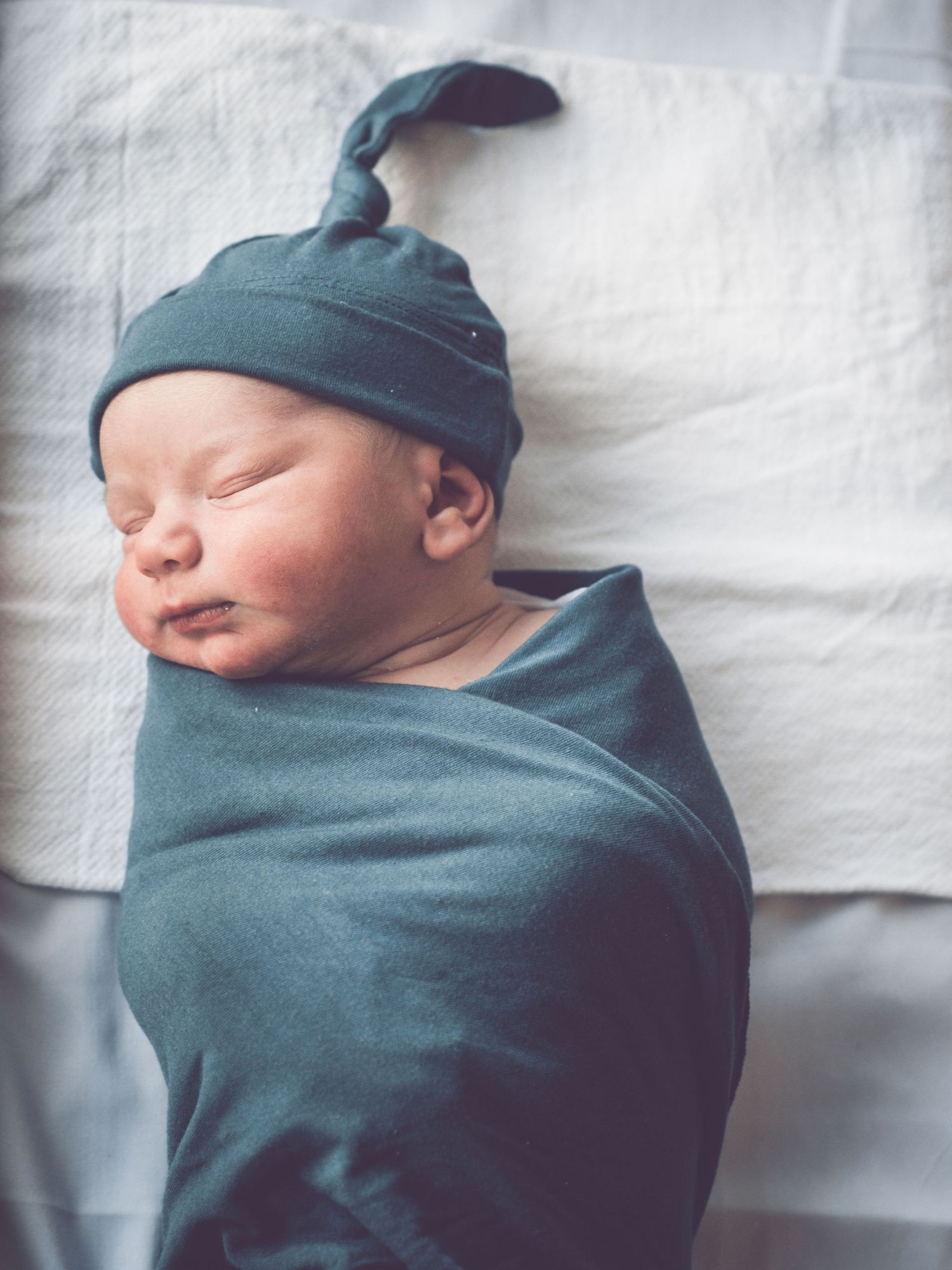 Sleeping newborn baby wrapped in teal swaddle wearing matching knotted hat.