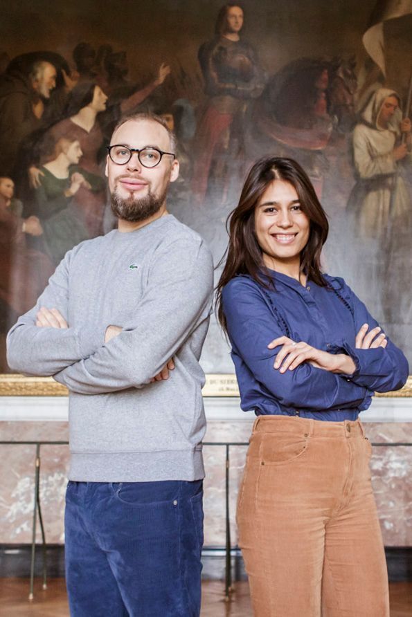 A couple poses together against a mural background wearing casual clothing in navy and gray with tan corduroy pants.