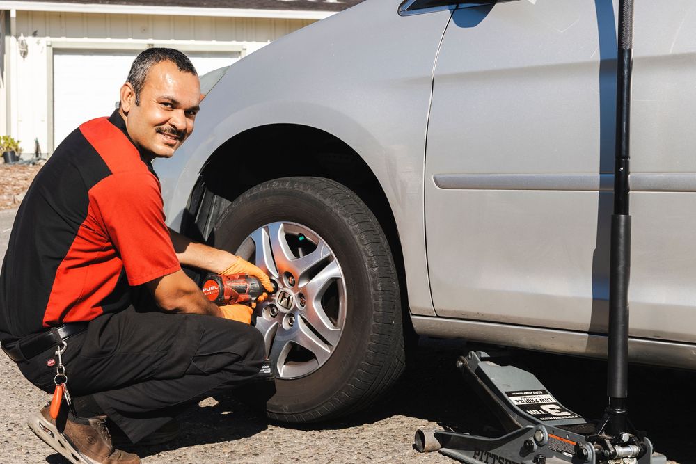 A mechanic in a red shirt working on a car's wheel while kneeling on the ground.